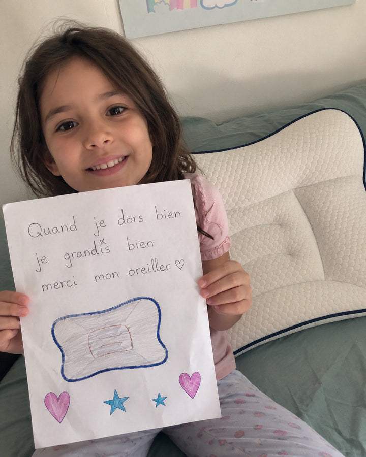 Petite fille souriante montrant un dessin fait à la main devant son oreiller ergonomique blanc à liseré bleu.