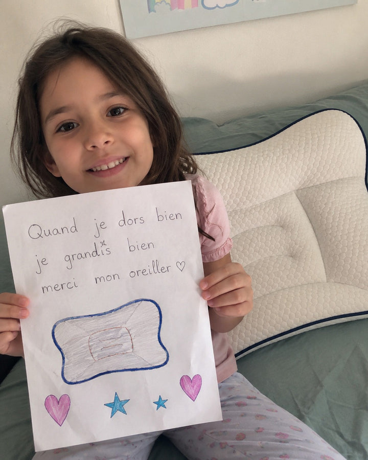 Petite fille souriante montrant un dessin fait à la main devant son oreiller ergonomique blanc à liseré bleu.